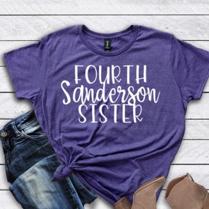 May include: Purple t-shirt with white text that reads "FOURTH Sanderson SISTER". The shirt is paired with blue jeans and brown ankle boots.