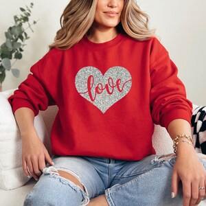 Puede incluir: Sudadera roja con un coraz&oacute;n de purpurina plateada que dice "love" en ella.