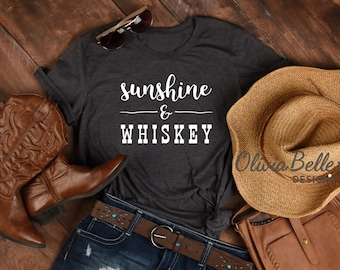 May include: A dark gray t-shirt with the text "sunshine & whiskey" in white lettering. The shirt is paired with denim shorts, brown cowboy boots, a straw hat, and a brown leather purse.