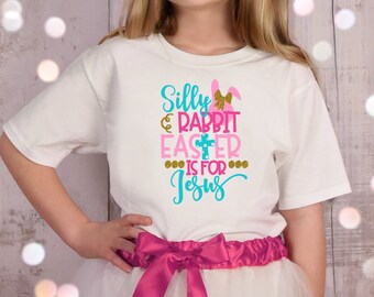 May include: A white t-shirt with a pink and blue graphic that says "Silly Rabbit Easter is for Jesus". The shirt is being worn by a child with a pink bow around their waist.