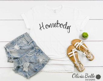 May include: White t-shirt with the text "Homebody" written in black cursive script. The shirt is laying on a white wooden surface with a pair of white sandals, a pair of denim shorts, and a small potted succulent.
