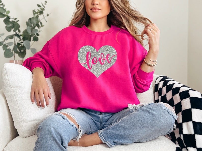 Puede incluir: Una sudadera rosa con un coraz&oacute;n de purpurina plateada que dice "love" en letra cursiva.