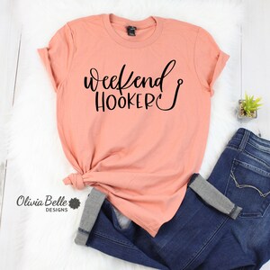 Peut inclure: Un t-shirt couleur p&ecirc;che avec le texte "weekend hooker" en &eacute;criture noire avec un symbole d'hame&ccedil;on de p&ecirc;che.
