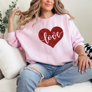 Puede incluir: Una sudadera rosa con un coraz&oacute;n rojo de purpurina que dice "love". La sudadera la lleva puesta una persona sentada en un sof&aacute; blanco.