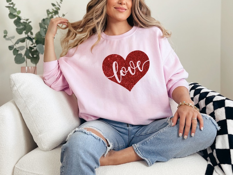Puede incluir: Una sudadera rosa con un coraz&oacute;n rojo de purpurina que dice "love". La sudadera la lleva puesta una persona sentada en un sof&aacute; blanco.