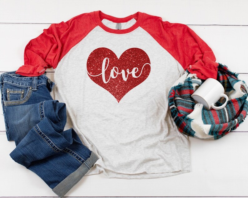 May include: A red and white long-sleeved baseball tee shirt with a red glitter heart that says "love" on it. The shirt is lying on a white wooden surface with blue jeans and a plaid scarf.