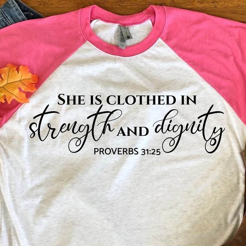 May include: A white and pink baseball tee shirt with the text "She is clothed in strength and dignity Proverbs 31:25" printed in black.