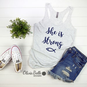 May include: A gray tank top with the text "She is strong" in blue lettering and a blue fish symbol. The tank top is laying on a white wooden surface with a pair of white sneakers and a pair of blue denim shorts.