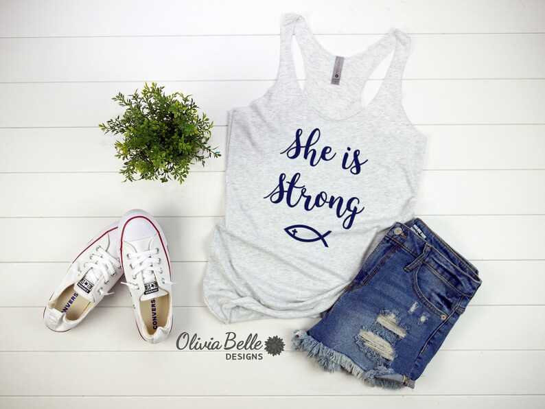 May include: A gray tank top with the text "She is strong" in blue lettering and a blue fish symbol. The tank top is laying on a white wooden surface with a pair of white sneakers and a pair of blue denim shorts.