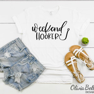 Peut inclure: T-shirt blanc avec un texte noir qui dit "Weekend Hooker" avec un symbole d'hame&ccedil;on de p&ecirc;che. Le t-shirt est pos&eacute; sur une surface en bois blanc avec une paire de sandales blanches et un short en jean.