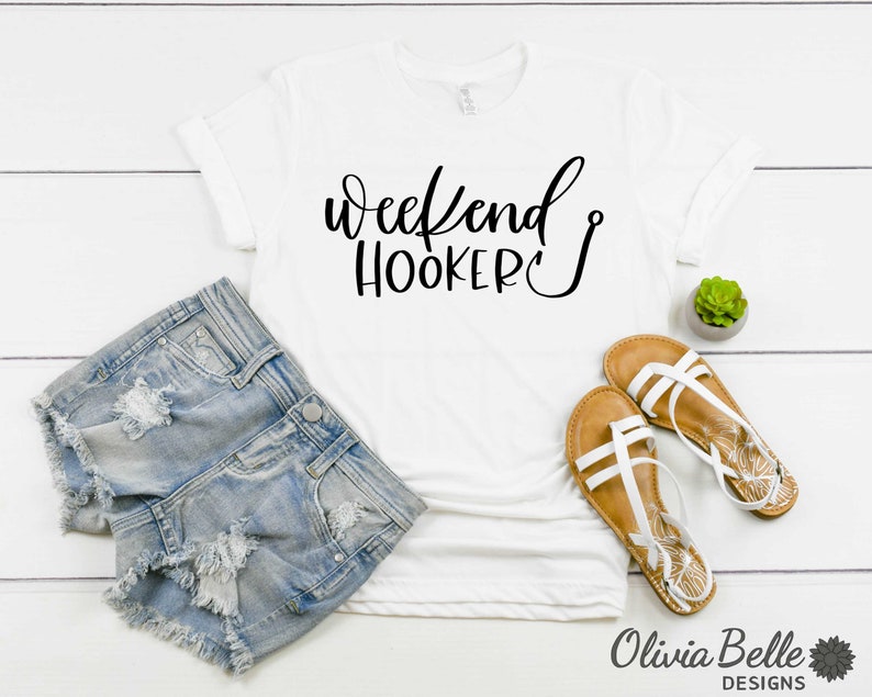Peut inclure: T-shirt blanc avec un texte noir qui dit "Weekend Hooker" avec un symbole d'hame&ccedil;on de p&ecirc;che. Le t-shirt est pos&eacute; sur une surface en bois blanc avec une paire de sandales blanches et un short en jean.