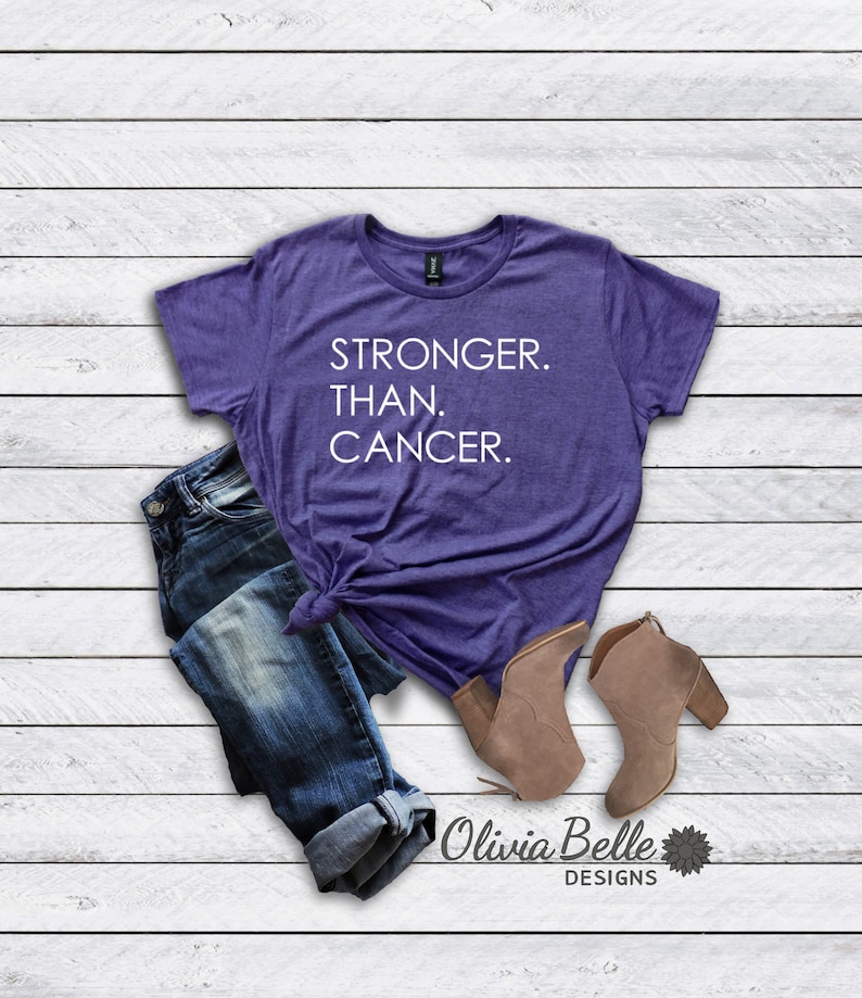 以下が含まれることがあります： 白い文字で「STRONGER. THAN. CANCER.」と書かれた紫色のTシャツ。Tシャツは青いジーンズと茶色のアンクルブーツとコーディネートされています。画像には、黒い文字で「Olivia Belle Designs」と小さな花のグラフィックが書かれています。