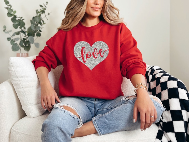 Puede incluir: Sudadera roja con un coraz&oacute;n de purpurina plateada que dice "love" en ella.