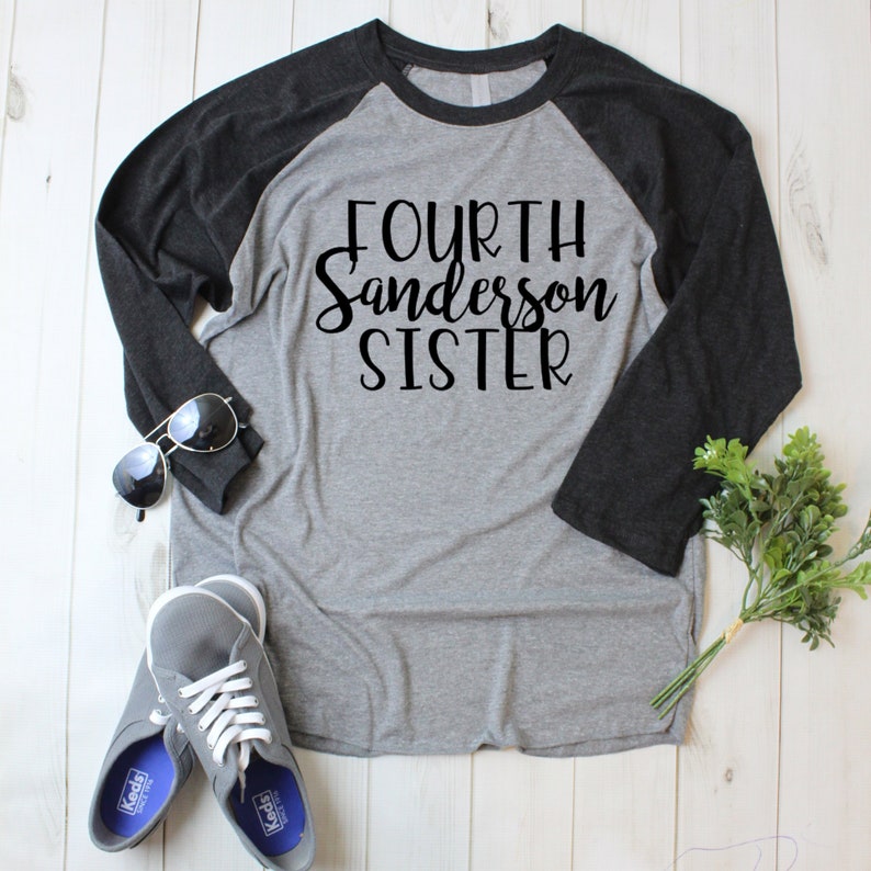 Puede incluir: Una camiseta de b&eacute;isbol gris y negra con el texto "FOURTH Sanderson SISTER" impreso en negro. La camiseta est&aacute; colocada sobre una superficie de madera blanca con unas gafas de sol y una peque&ntilde;a planta.