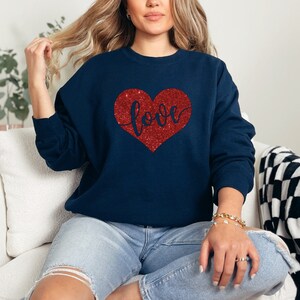 Puede incluir: Una sudadera azul marino con un coraz&oacute;n rojo de purpurina que dice "love" en letra cursiva.