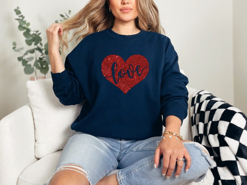 Puede incluir: Una sudadera azul marino con un coraz&oacute;n rojo de purpurina que dice "love" en letra cursiva.