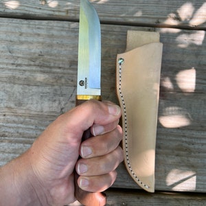 May include: A Skogsman knife with a wooden handle and a silver blade, held in a hand. The knife has a brass bolster and is accompanied by a light brown leather sheath. The sheath has white and black stitching along the edge.