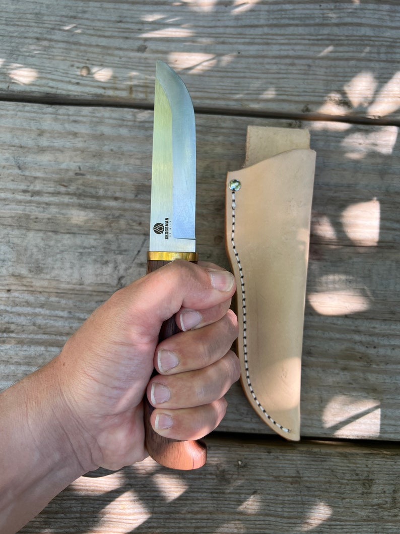 May include: A Skogsman knife with a wooden handle and a silver blade, held in a hand. The knife has a brass bolster and is accompanied by a light brown leather sheath. The sheath has white and black stitching along the edge.