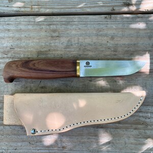May include: A Skogsmann knife with a wooden handle and a stainless steel blade, resting on a wooden surface. The knife is accompanied by a light tan leather sheath. The knife's handle is a dark brown color, and the blade is silver.