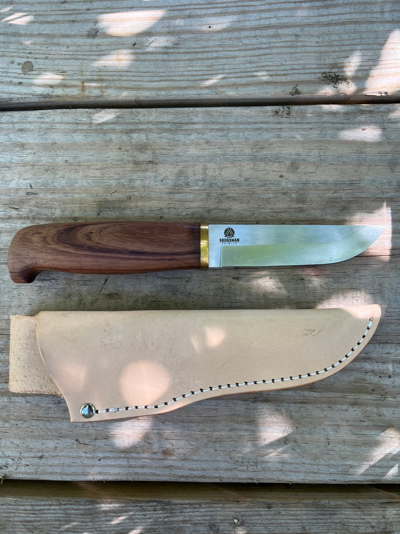 May include: A Skogsmann knife with a wooden handle and a stainless steel blade, resting on a wooden surface. The knife is accompanied by a light tan leather sheath. The knife's handle is a dark brown color, and the blade is silver.