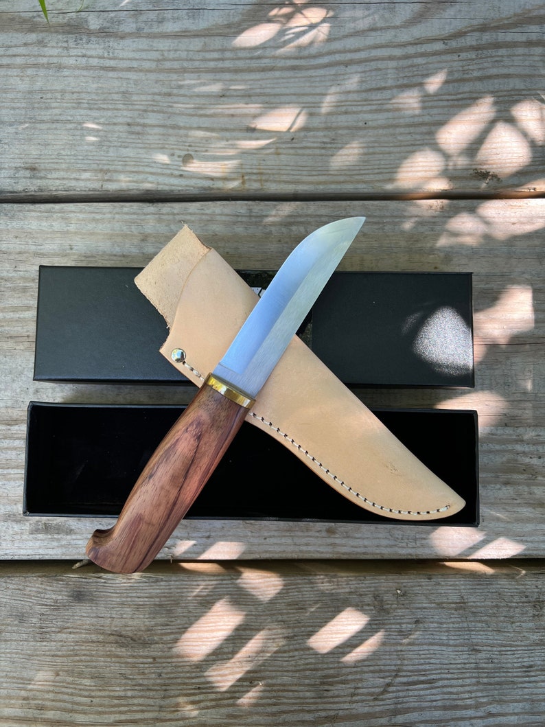 May include: A stainless steel knife with a wooden handle and a leather sheath. The knife has a curved blade and a brass bolster. The sheath is light brown and stitched with white thread. The knife and sheath are in a black box.