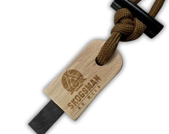 May include: A wooden fire starter with a black striker and brown cord. The wooden piece has the text "SKOGSMAN" and a logo etched into it. The cord is knotted and attached to a black handle.