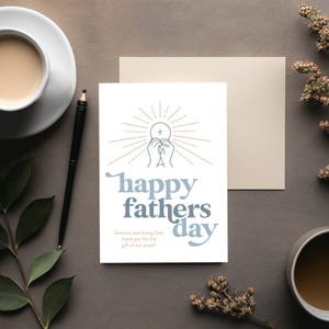 A white greeting card with "happy fathers day" and a communion host graphic is on a dark gray surface, with a pen, coffee, and dried flowers.