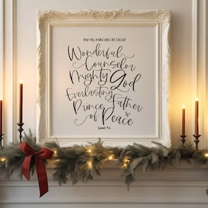 May include: A white framed print with a black calligraphy quote about Jesus. The quote reads "And his name will be called Wonderful Counselor Mighty God Everlasting Father Prince of Peace" with Isaiah 9:6 at the bottom. The print is displayed on a white mantel with a green garland and red bow.