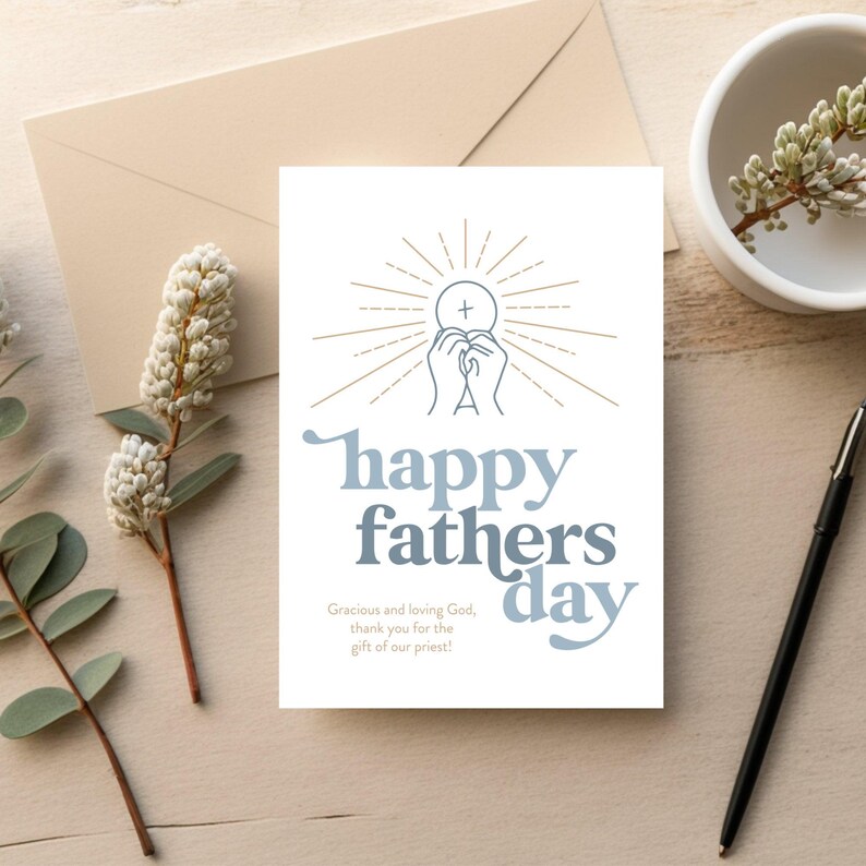 A white greeting card with "happy fathers day" and a communion host graphic is on a light wood surface next to a pen, a brown envelope, and a white floral sprig.