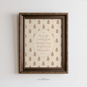 May include: A framed wall art print featuring a cream background with a brown diamond grid pattern and small floral motifs. An oval center contains the text: Don't judge each day by the harvest you reap, but by the seeds that you plant.