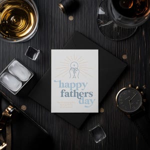 A white greeting card with "happy fathers day" and a communion host graphic is on a black envelope, surrounded by ice cubes, drinks, and a watch on a dark wood table.