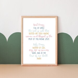 May include: A framed print with a light brown frame and a white background. The print features the Hail Mary prayer in a colorful handwritten font.