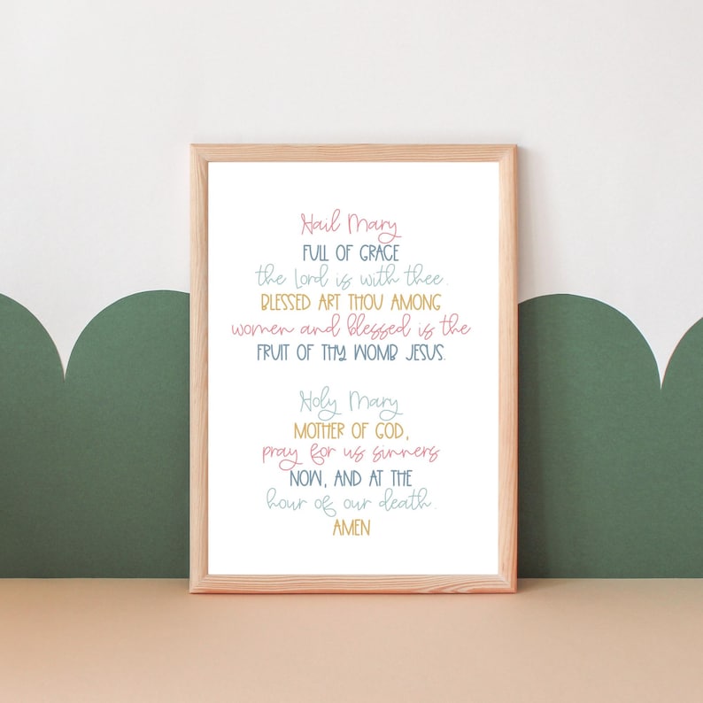 May include: A framed print with a light brown frame and a white background. The print features the Hail Mary prayer in a colorful handwritten font.