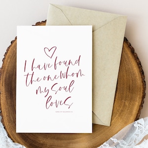 May include: A white greeting card with the text "I have found the one whom my soul loves" in burgundy script, with a heart above the text. A kraft paper envelope rests beside the card on a wooden surface.