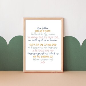 May include: A framed print with a white background and a light brown frame. The print features the Lord's Prayer in a handwritten style with blue, pink, and yellow text.