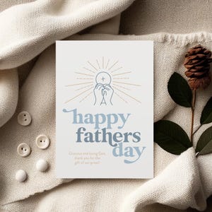 A white greeting card with "happy fathers day" and a communion host graphic is on a textured cream blanket, with white buttons and a pinecone.