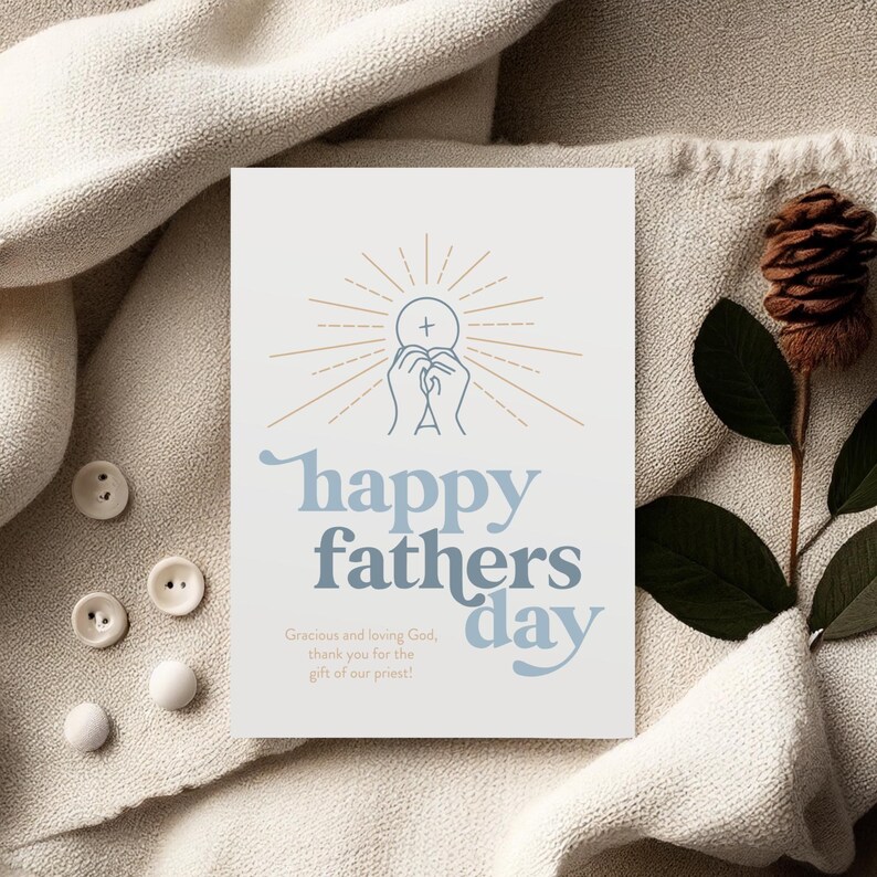 A white greeting card with "happy fathers day" and a communion host graphic is on a textured cream blanket, with white buttons and a pinecone.