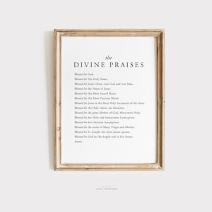 May include: A framed print featuring the text "the DIVINE PRAISES" in a minimalist font. The text is set against a white backdrop, and the frame is a light brown. The print contains a list of religious blessings.