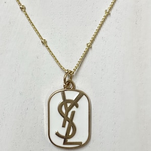 May include: Gold-toned necklace featuring a rectangular pendant with a white enamel background and a gold logo. The chain is delicate with small gold beads. The pendant is approximately 1 inch in length.
