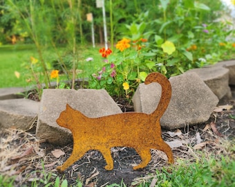 May include: A rusty orange metal cat silhouette walking on grass. The cat has a curled tail and is placed in front of a garden bed with flowers and concrete blocks.