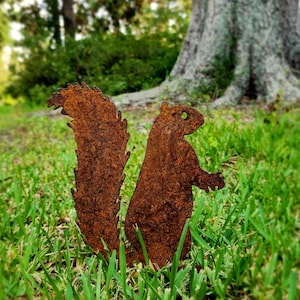 May include: A rusty metal squirrel silhouette stands in green grass. The squirrel has a large, textured tail and is posed with its arms out. The background includes a blurred tree and foliage, suggesting an outdoor setting.