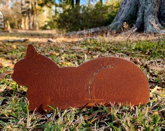 May include: A rusty brown metal cat silhouette, lying down in a grassy area. The cat's tail is curled, and the metal has a textured, weathered appearance. The cat is a decorative garden accent.