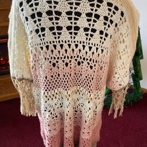 May include: A handmade, open-weave crochet cardigan in a gradient of colors. The top is cream, transitioning to light pink, then to a darker pink and tan at the bottom. The sleeves have fringe at the cuffs. The back features a geometric openwork design.
