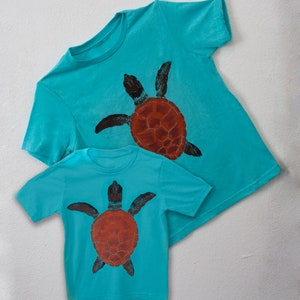 May include: Two turquoise t-shirts with a sea turtle graphic printed on the front. The larger shirt has a larger turtle graphic and the smaller shirt has a smaller turtle graphic.