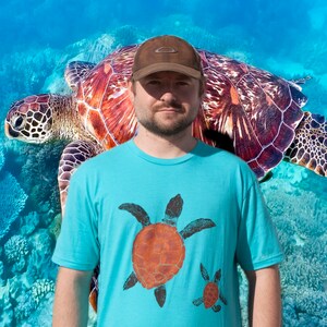 May include: A light blue t-shirt featuring two sea turtle designs in orange and black. The person in the image is wearing a brown baseball cap. A large sea turtle is in the background, with a blue and white coral reef backdrop.