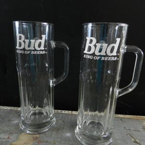 May include: Two clear glass Budweiser beer mugs with handles. The mugs have the Budweiser logo and the text "King of Beers" printed on them.