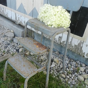 Puede incluir: Un taburete de metal gris envejecido con un pequeño respaldo, con dos escalones y un asiento. Un gran racimo de flores blancas descansa sobre el asiento. El taburete muestra signos de óxido y desgaste, sobre un fondo de pared y grava desgastadas.