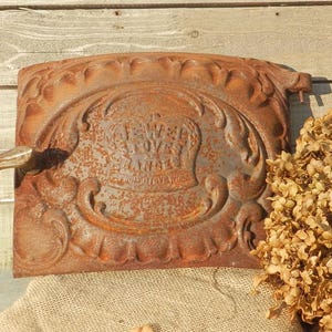 Puede incluir: Puerta de estufa antigua de hierro fundido con adornos de volutas y el texto "JEWEL STOVES RANGES". La puerta, con un asa, está muy oxidada, mostrando su antigüedad. Un arreglo floral seco está al lado.