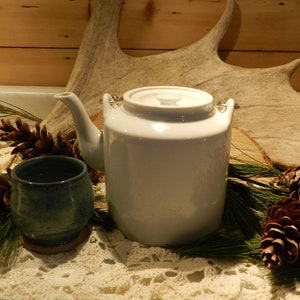 May include: A white ceramic teapot with a lid and a handle, sitting on a white crocheted tablecloth with pine branches and pine cones.
