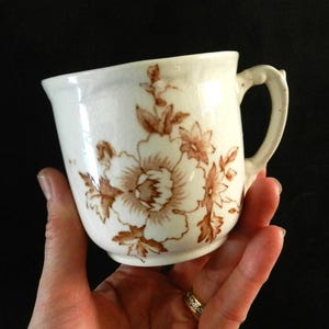 May include: Cream-coloured ceramic mug with a brown floral pattern. The mug has a curved handle and a slightly angled shape. The floral design features large, stylised flowers and leaves.